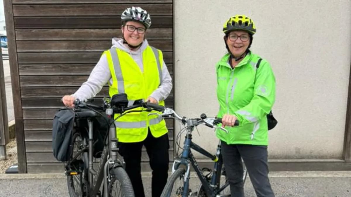Two women wearing cycling helmets and reflective jackets stand smiling next to their bicycles.