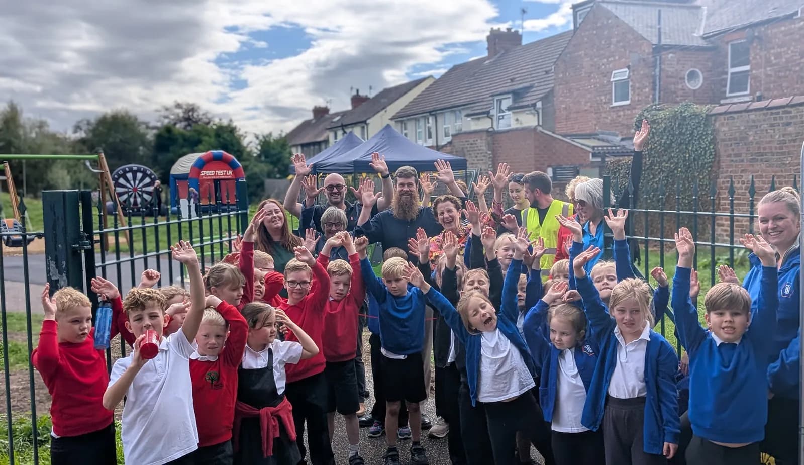 A large group of children and adults, mostly smiling, stand in a line behind a black metal fence, all waving their hands in the air, with a residential street and a small park visible in the background.