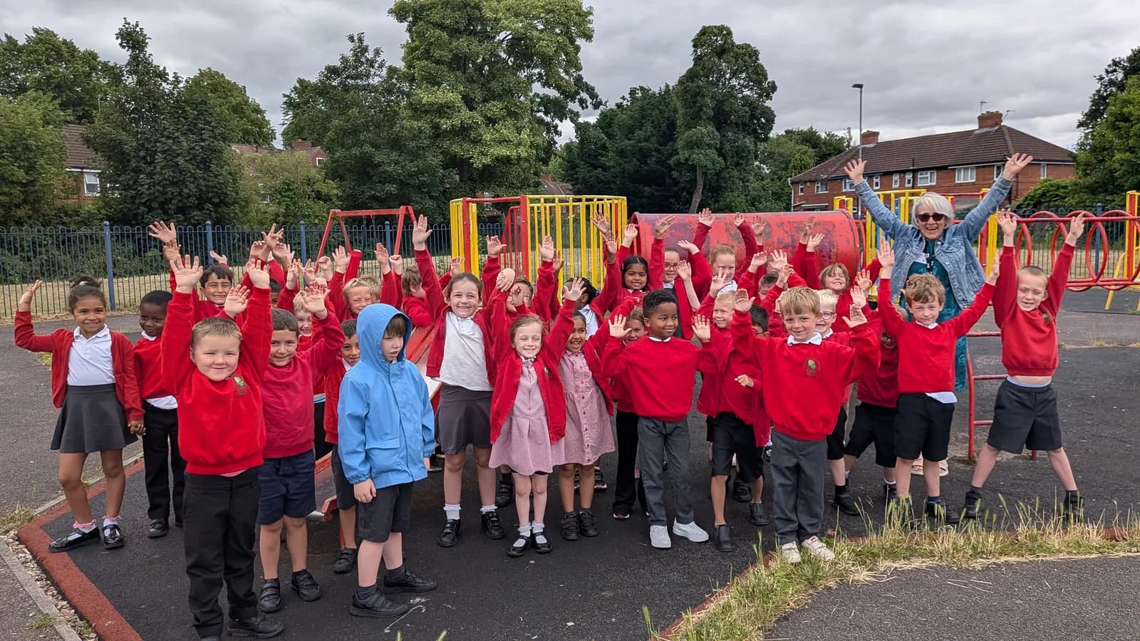 A group of elementary school children in red uniforms and one adult in a denim jacket joyfully raise their hands in a playground setting.