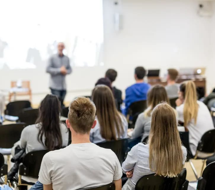A man in a light blue shirt stands and talks in front of a projector screen to a group of students sitting with their backs to the viewer.