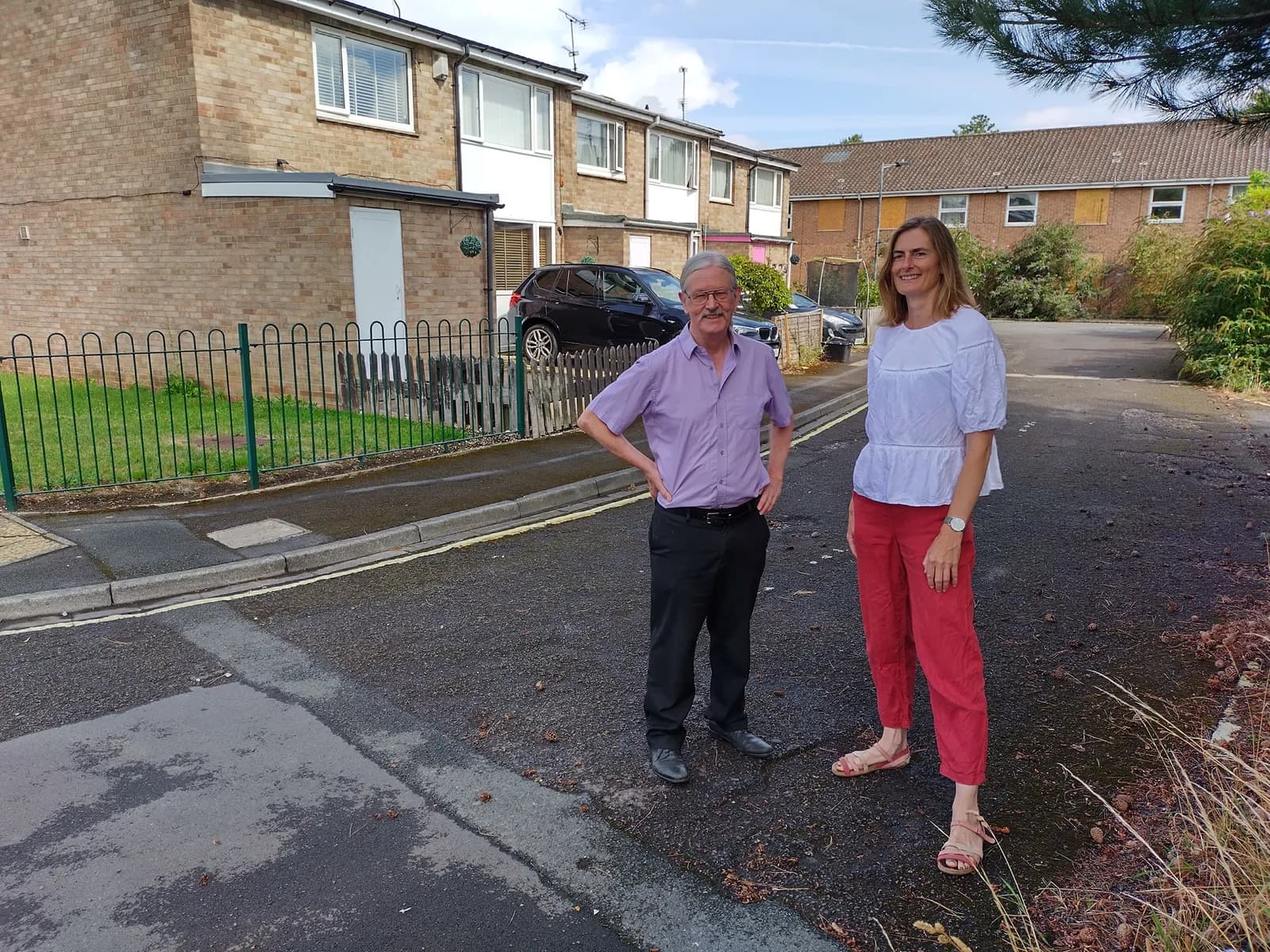 A man and a woman stand on a paved area in front of residential buildings, with a dark car parked in the background and a fence to their left.