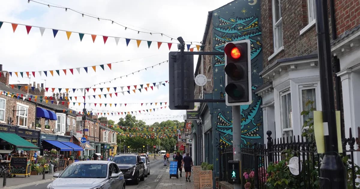 A street view of a small town with colorful bunting strung across the road, cars parked and driving, and brick buildings with storefronts.