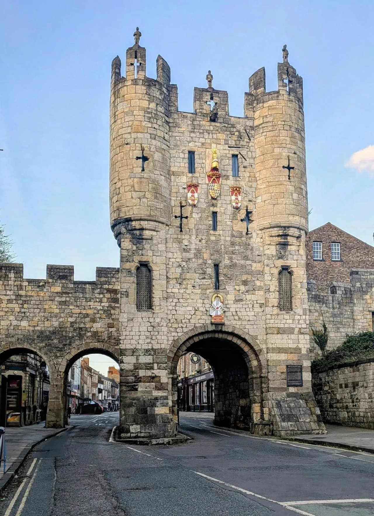 A historic stone gatehouse with two towers and arched passages spanning a street, adorned with coats of arms and a blue sky above.