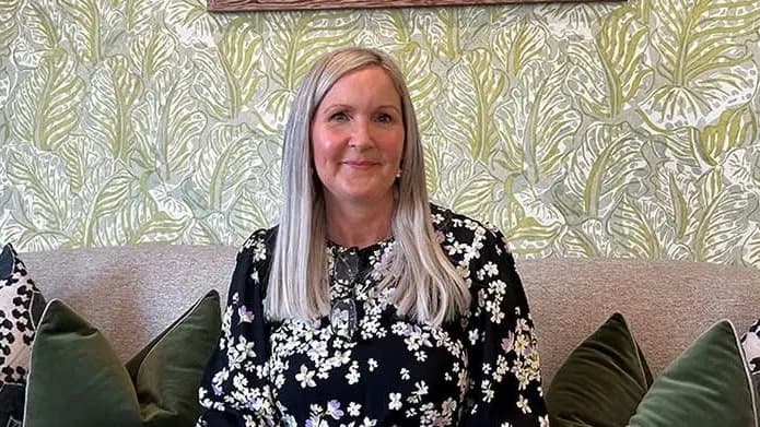 A smiling woman with long grey hair and a black floral dress sits on a couch in front of green leaf-patterned wallpaper.